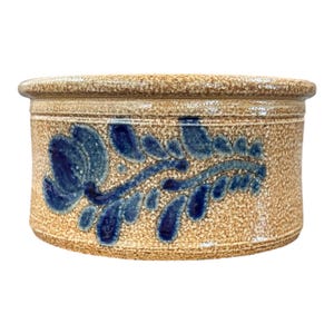 May include: A cylindrical stoneware crock featuring a speckled tan and beige finish with a prominent blue hand-painted floral motif. The vessel has a thick, rounded rim and a smooth, glazed texture, showcasing a classic design with a wide opening.