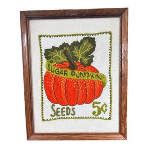 May include: A framed needlepoint art piece featuring an orange pumpkin with green leaves and a yellow banner reading SUGAR PUMPKIN. Below the pumpkin, the text reads SEEDS 5 cents. The embroidery is set on a light fabric inside a brown wood frame.