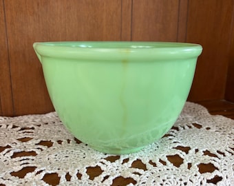 May include: A light green jadeite mixing bowl with a smooth, rounded rim and a small pouring spout on the side. The bowl features a translucent, glass-like finish with a subtle vertical brown discoloration mark running down the front surface.