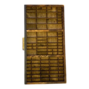 May include: A vintage wooden printer's tray with numerous small compartments of varying sizes. The tray is rectangular, with a dark brown frame and a lighter, aged wood interior. A small, gold-coloured metal clasp is visible on the left side.