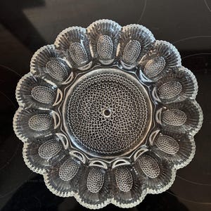 Op de afbeelding: Een heldere glazen serveerschaal voor gevulde eieren. De schaal heeft een cirkelvormig midden met een patroon van kleine cirkels en is omgeven door 12 eivormige inkepingen. De buitenrand heeft een geschulpte vormgeving.