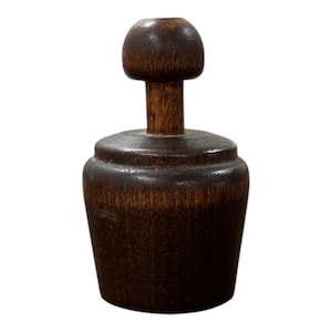 May include: A dark brown wooden butter stamp featuring a cylindrical base with a stepped top and a central vertical handle topped with a rounded knob. The wood grain is visible throughout the surface of the object, showing a smooth, polished finish.