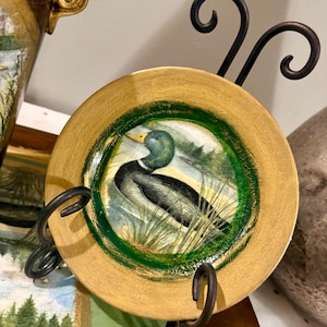 May include: A decorative plate featuring a painted duck scene, set within a green border and a gold-colored rim. The plate is displayed on a black metal stand, showcasing a nature-inspired design. The plate is round.