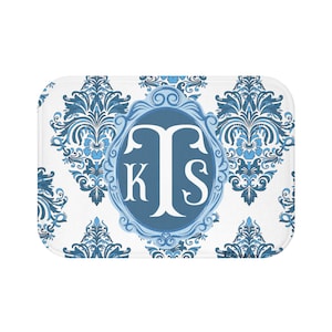Puede incluir: Alfombra de baño rectangular blanca con un patrón de damasco azul y un diseño ovalado central. El óvalo contiene las letras blancas "KTS" con un borde azul. La alfombra tiene bordes redondeados.