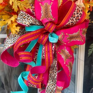 Peut inclure: Une couronne décorative composée de feuilles de chêne aux couleurs automnales et d'un grand nœud multicouche. Le nœud est réalisé avec des rubans rose vif, turquoise et orange, ornés de motifs léopard et d'illustrations de guépards.