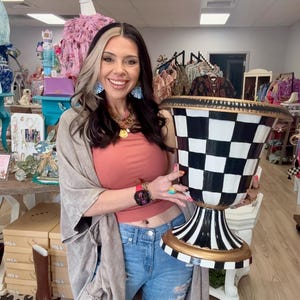 May include: A large, decorative urn with a black and white checkered pattern, gold trim, and a pedestal base. The urn is held by a person in a retail setting, showcasing its size and design.