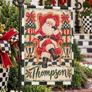 May include: A rectangular garden flag featuring a painted illustration of Santa Claus sitting in a red chair. The border has a black and white checkered pattern. The name Thompson is written in green script at the bottom with holly accents.