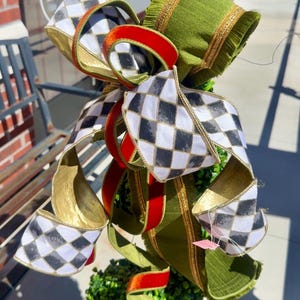 May include: A decorative bow featuring a black and white diamond harlequin pattern, solid red velvet ribbon, and olive green fabric with gold trim. The bow is attached to a topiary made of small green leaves, creating a layered, textured appearance.