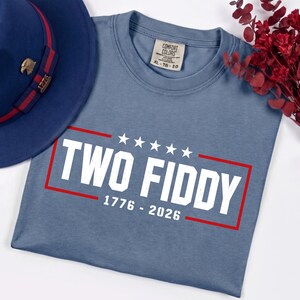 May include: A blue crew neck t-shirt featuring the white text TWO FIDDY, five white stars, and the numbers 1776 - 2026 enclosed in a red rectangular border. The shirt has a Comfort Colors label showing size XL, and is next to a blue fedora hat.
