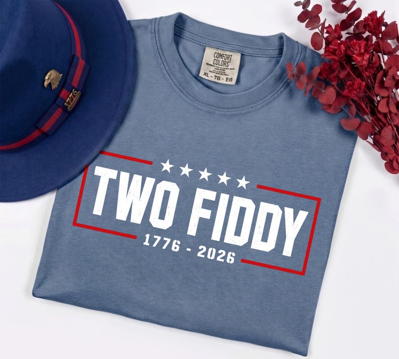 May include: A blue crew neck t-shirt featuring the white text TWO FIDDY, five white stars, and the numbers 1776 - 2026 enclosed in a red rectangular border. The shirt has a Comfort Colors label showing size XL, and is next to a blue fedora hat.