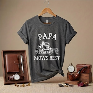 May include: Dark gray t-shirt with the text "PAPA MOWS BEST" and a graphic of a riding lawn mower. The shirt is displayed on a wooden hanger. Other items include a watch, wallet, pen, pipe, and pocket watch.