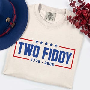 May include: A cream-colored crewneck t-shirt featuring a blue and red graphic that reads TWO FIDDY 1776 - 2026 with five blue stars above the text. A navy blue fedora with a red and blue striped band and a gold eagle pin sits to the left.