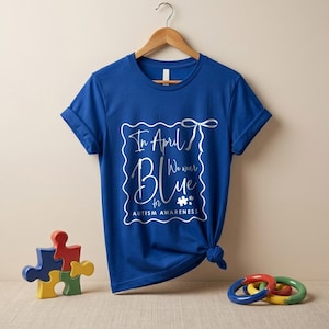 May include: A royal blue t-shirt with the text "In April We Wear Blue for Autism Awareness" in a white, decorative frame. The shirt is knotted at the side and hangs on a wooden hanger.