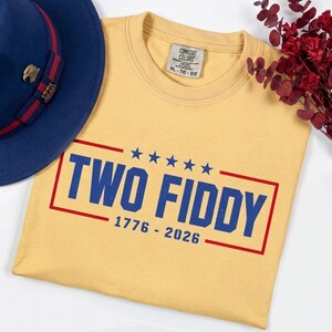 May include: A yellow t-shirt with a blue and red graphic print that reads TWO FIDDY 1776 - 2026 with five blue stars above the text. A blue hat with a red band and a gold eagle pin is positioned to the left of the shirt, and red dried flowers.