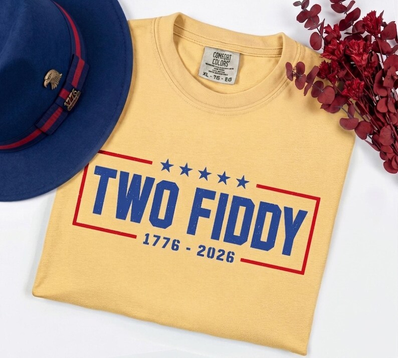May include: A yellow t-shirt with a blue and red graphic print that reads TWO FIDDY 1776 - 2026 with five blue stars above the text. A blue hat with a red band and a gold eagle pin is positioned to the left of the shirt, and red dried flowers.