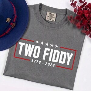 May include: A grey t-shirt featuring the white text TWO FIDDY with five stars above and the years 1776 - 2026 below, all enclosed in a red rectangular border. The shirt has a Comfort Colors label at the neck and is displayed with a blue hat.