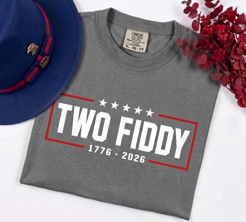 May include: A grey t-shirt featuring the white text TWO FIDDY with five stars above and the years 1776 - 2026 below, all enclosed in a red rectangular border. The shirt has a Comfort Colors label at the neck and is displayed with a blue hat.