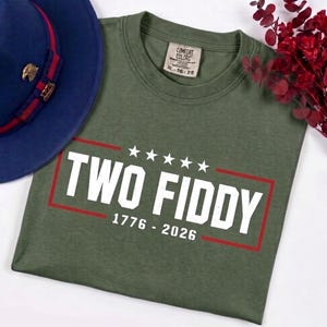 May include: An olive green t-shirt featuring the white text TWO FIDDY, five white stars, and the years 1776 - 2026 enclosed in a red rectangular border. The shirt has a Comfort Colors label indicating size XL and is paired with a blue fedora hat.
