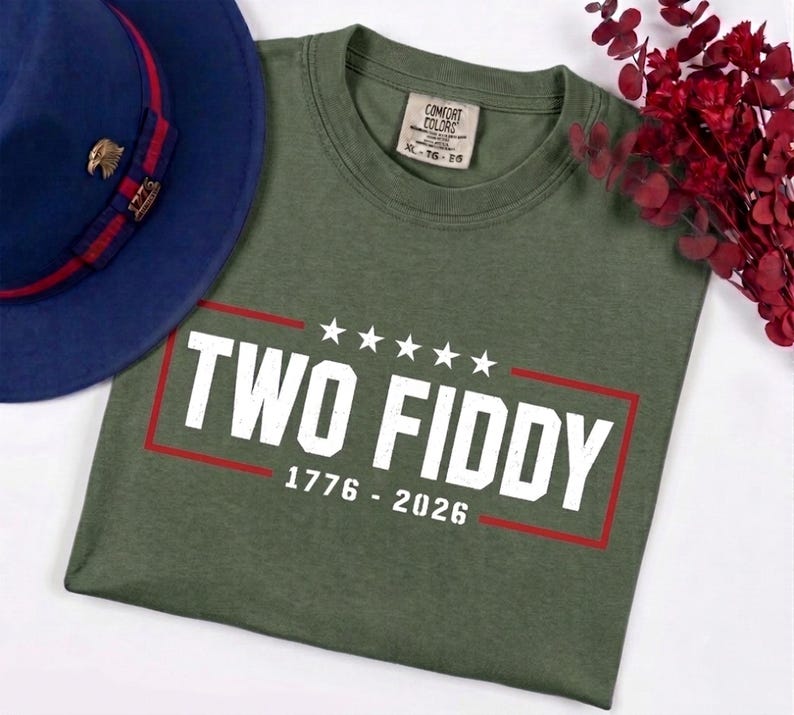 May include: An olive green t-shirt featuring the white text TWO FIDDY, five white stars, and the years 1776 - 2026 enclosed in a red rectangular border. The shirt has a Comfort Colors label indicating size XL and is paired with a blue fedora hat.