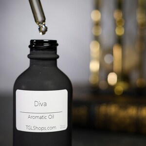 May include: A black bottle of Diva Aromatic Oil with a white label, a glass dropper dispensing oil, and a blurred background. The label includes the website TGLShops.com. The bottle is 2 oz.