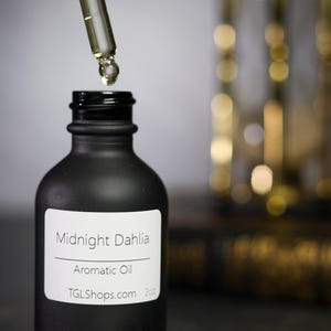 May include: A black matte bottle of Midnight Dahlia Aromatic Oil with a dropper dispensing liquid. The bottle has a white label with the product name and website. A blurred hourglass is in the background.