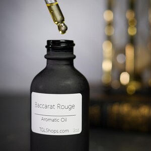May include: A black matte bottle of Baccarat Rouge aromatic oil with a dropper dispensing the oil. The label reads "Baccarat Rouge Aromatic Oil TGLShops.com 2oz."