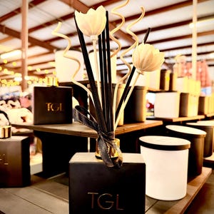 May include: Black square diffuser bottle with gold accents and the letters "TGL". The diffuser has black reeds, white flower-shaped decorations, and curly accents. A black ribbon is tied around the neck. Other black and white candles are in the background.