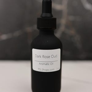 May include: A matte black glass bottle with a dropper, labeled "Dark Rose Oud Aromatic Oil." The bottle has a black cap and a white label with the website TGLShops.com. The background is a dark, textured surface.