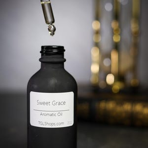 May include: A black bottle of aromatic oil with a white label that reads "Sweet Grace." A glass dropper dispenses the oil into the bottle. The website TGLShops.com is also visible on the label.