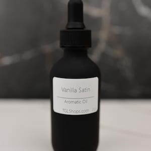 May include: A black matte bottle with a dropper top, labeled "Vanilla Satin Aromatic Oil." The label also includes the website TGLShops.com. The bottle is set against a dark background.