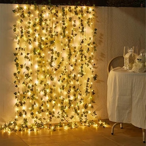 May include: A wall decorated with hanging faux ivy and warm white fairy lights. The lights are strung vertically, creating a glowing backdrop. A table with a white tablecloth and decorative items is to the right.