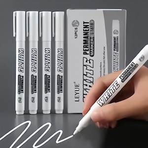 May include: White permanent markers with the text "WHITE PERMANENT" on the barrel and a box of 12 markers. One marker is being used to draw a white line on a dark surface. The brand name "LEYUE" is visible on the box.