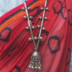 May include: Silver-toned necklace with a black cord. The necklace features a series of small, round beads and a decorative pendant with dangling beads. The background includes a red fabric with dark blue circular patterns.