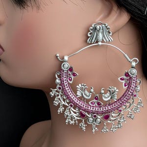 May include: A large silver hoop earring featuring an oxidized finish, adorned with small pink stones and intricate metalwork. The design includes two central bird figures, decorative floral motifs, and dangling silver beads along the bottom edge.