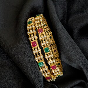 May include: Two gold-toned bangles featuring a repeating pattern of square-cut red and green stones. The metal frame is decorated with small white beads arranged in rows between the colored stones, creating a textured and detailed circular design.