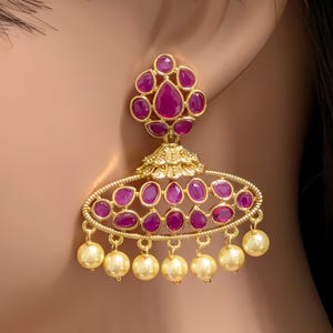 May include: A gold-toned jhumka earring featuring a floral-shaped stud set with deep pink teardrop stones. The bell-shaped component is adorned with an oval frame of matching pink stones, finished with a row of dangling cream-colored pearl beads.