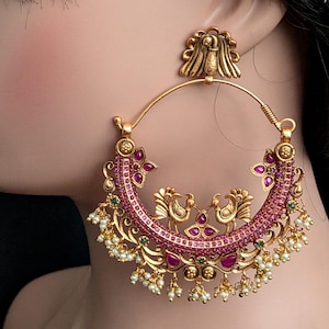 Peut inclure: Une grande boucle d'oreille créole dorée avec un clou en forme de paon. La base en croissant est ornée de petites pierres roses, de touches vertes et de deux paons centraux, le tout agrémenté de perles de rocaille blanches suspendues.