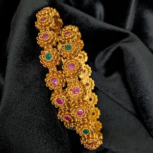 May include: Two gold-toned bangles featuring a repeating floral motif. Each flower is textured with a matte finish and centered with either a small round pink or green gemstone. The bangles have a continuous circular design with intricate metalwork.