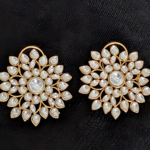May include: A pair of gold-toned stud earrings. Each earring features a central, round, clear stone surrounded by numerous pear-shaped, clear stones. The earrings have a floral design and are set against a black background.