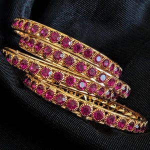 May include: A set of three gold-toned bangles, each featuring a continuous row of circular, deep pink faceted stones set in a scalloped metal frame. The bracelets are arranged in a stacked formation, showcasing the metallic luster and stones.