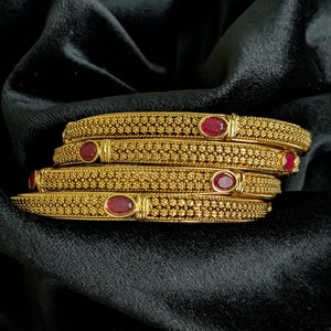 May include: A set of four gold-toned bangles featuring a textured, granular surface and inlaid with small, oval-shaped red stones. Each bangle displays a repeating pattern of tiny raised dots across the band, with the red stones set at intervals.