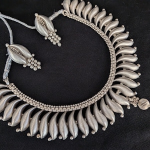 May include: A silver-toned necklace and matching earrings set. The necklace features a series of leaf-shaped pendants and a delicate chain. The earrings have a similar design. The jewelry is displayed against a black background.