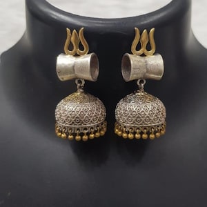 May include: A pair of dangling earrings featuring a gold-toned trident symbol at the top, a silver-toned cylindrical connector, and a bell-shaped jhumka base with intricate silver filigree patterns and small gold-toned beads hanging from the bottom edge.
