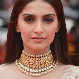 Op de afbeelding: Een vrouw draagt een gouden choker-ketting met parels en roze edelstenen. De ketting is gelaagd en versierd. Ze draagt een lichtgekleurde, geborduurde top. De tekst onderaan de afbeelding luidt "FOR REFERENCE PURPOSE ONLY."