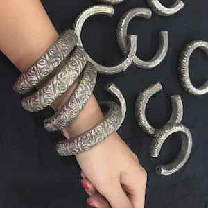 Può includere: Diversi braccialetti aperti color argento con un motivo ornamentale ripetuto. Alcuni braccialetti sono impilati su un polso, altri sono sparsi. I gioielli hanno un'estetica vintage o antica. Probabilmente in metallo.