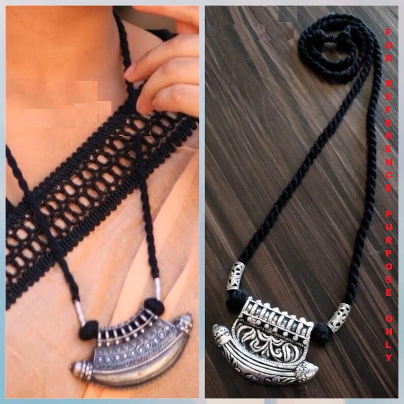 May include: A silver-toned pendant necklace with intricate detailing and a black cord. The pendant has a curved, crescent-like shape with ornate patterns. The necklace is displayed on a light-colored fabric and a wooden surface.