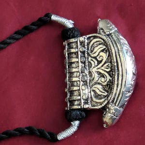 May include: Silver-toned pendant necklace with intricate floral and geometric designs. The pendant is attached to a black cord necklace. The background is a solid maroon color.