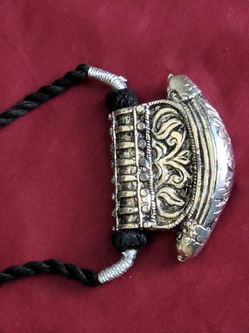 May include: Silver-toned pendant necklace with intricate floral and geometric designs. The pendant is attached to a black cord necklace. The background is a solid maroon color.