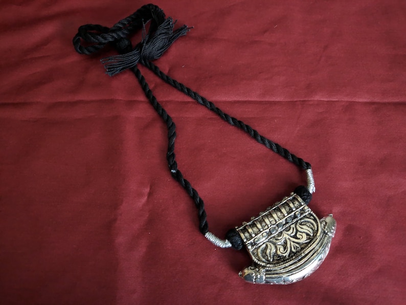 May include: An ornate silver and gold-toned pendant necklace with a black rope cord. The pendant features intricate floral designs and is suspended from a twisted black cord with tassels. The necklace is displayed on a red background.