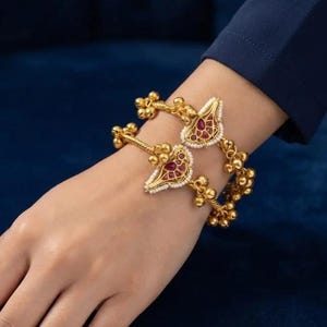 May include: Two gold-toned bangles featuring a central teardrop-shaped motif inlaid with red stones and white beads. The bangles are adorned with multiple small gold-toned spherical charms attached along the band, creating a textured appearance.